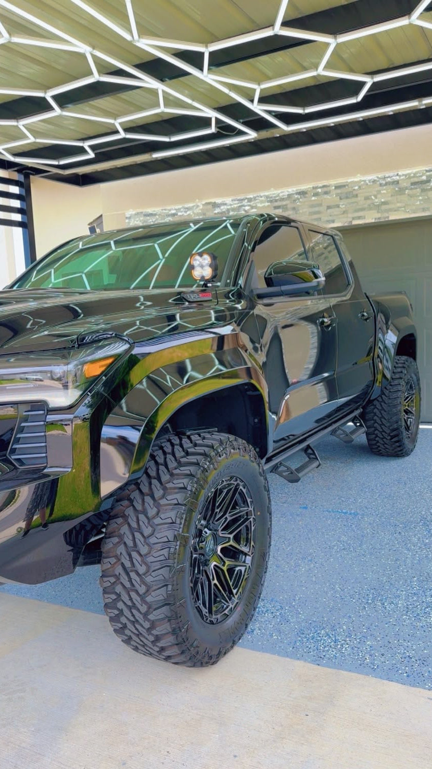 A glossy black lifted pickup truck with large off-road tires and black wheels, parked inside a modern garage with a geometric ceiling light fixture and blue-gray concrete floor.